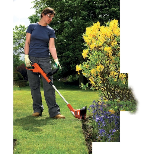 A person uses a BLACK+DECKER cordless grass trimmer to edge a lawn next to a flowerbed with yellow and purple flowers. The person is wearing gloves and outdoor clothing, trimming the grass in a garden.