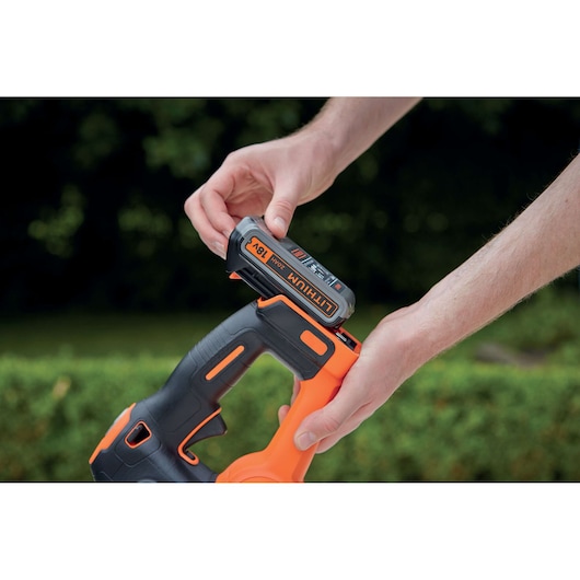 Close-up view of a person inserting an 18V lithium battery into a BLACK+DECKER cordless power tool outdoors, with greenery in the background.