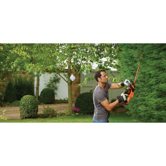 A person using an orange and black BLACK+DECKER cordless hedge trimmer to cut green hedges in a garden. The person is wearing gloves and a grey t-shirt.