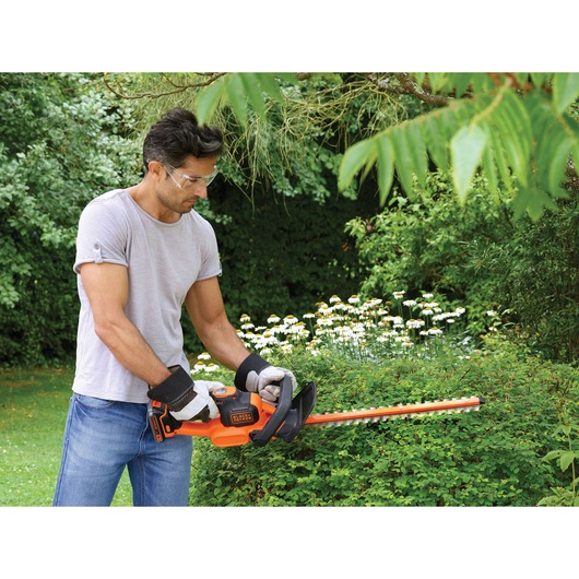 A person with a blurred face is using a BLACK+DECKER cordless hedge trimmer to trim bushes in a garden. The trimmer is orange and black, and the person is wearing gloves and a grey T-shirt.