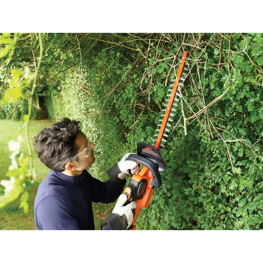A person with a blurred face uses a BLACK+DECKER cordless hedge trimmer to cut branches and leaves from a dense green hedge in a garden.