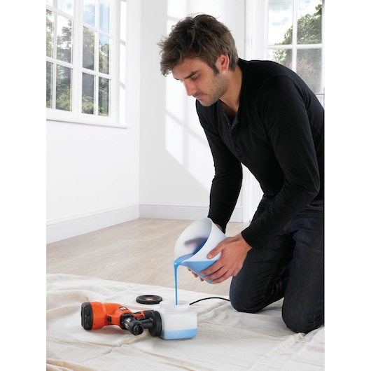 A person kneeling indoors pours blue liquid from a bowl into the container of an orange BLACK+DECKER HVLP200 paint sprayer, which is placed on a cloth-covered floor.
