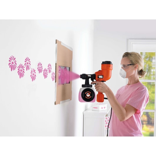 A person uses a BLACK+DECKER HVLP200 paint sprayer to apply pink flower patterns on a white wall through a stencil.