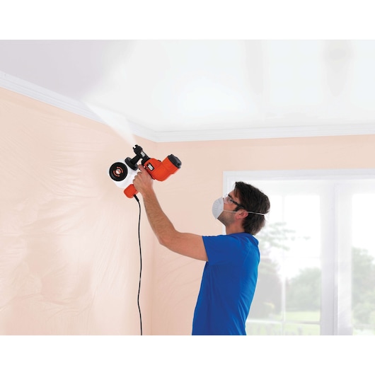A person with a blurred face uses a BLACK+DECKER HVLP200 paint sprayer to spray white paint onto a ceiling indoors. The person is wearing a blue shirt and stands in a room with light peach walls and a window in the background.
