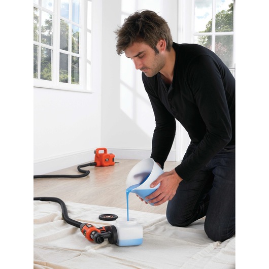 A person kneeling on a sheet indoors, pouring blue liquid into the container of a BLACK+DECKER HVLP400 paint sprayer. An orange paint sprayer unit and hose are visible on the floor, with large windows in the background.