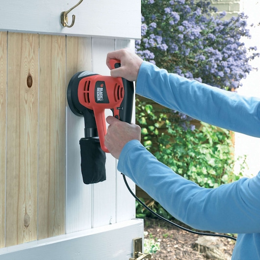 A person is using a BLACK+DECKER KA191EK electric sander on a wooden door. The sander is red and black, corded, and being used to smooth the wood surface.