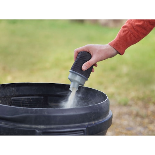 A hand is holding a small cylindrical filter attachment and releasing fine dust into a black container outdoors.