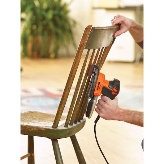 A person using a BLACK+DECKER KA2500K electric sander to sand the slats of a wooden chair indoors.