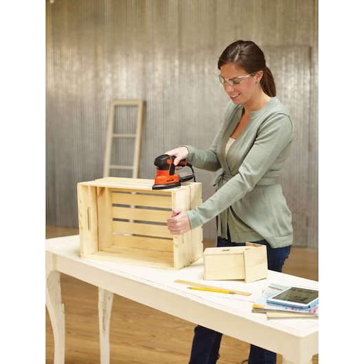 A person sanding a wooden crate with a BLACK+DECKER electric sander on a white table. Other wooden boxes, a ruler, pencil, notebook, and tablet are also on the table. The person's face is blurred.