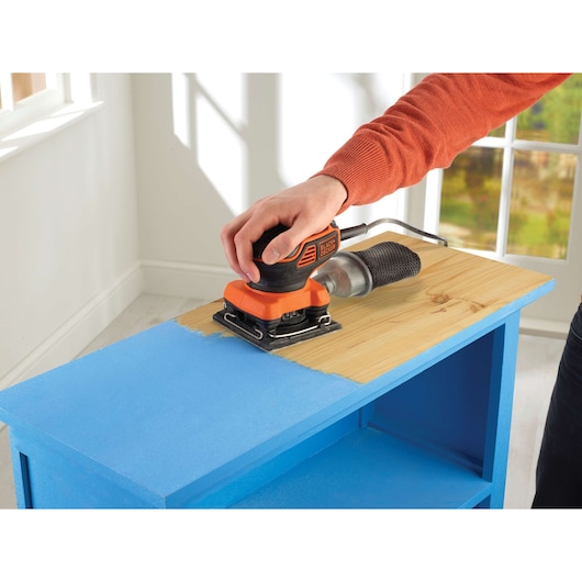 A person uses a BLACK+DECKER KA450 electric sander to remove blue paint from the top surface of a wooden shelf in a bright room.