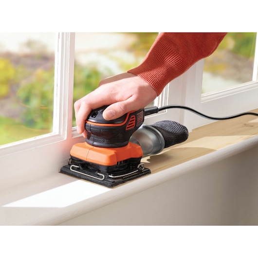 A close-up of a person's hand using a BLACK+DECKER KA450 electric sander to smooth a wooden window sill indoors.
