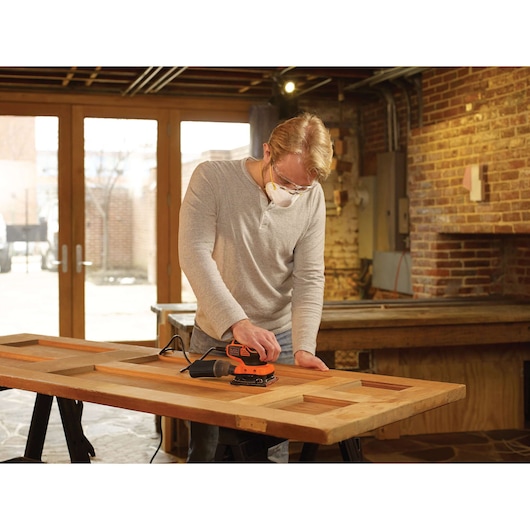 A person using a BLACK+DECKER KA450 electric sander to sand a wooden door on a workbench in a workshop.