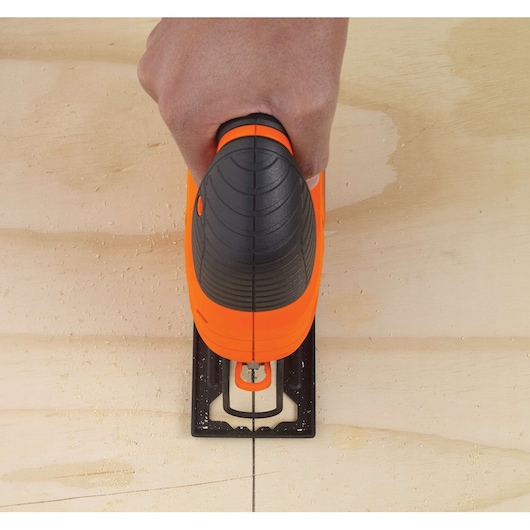 A hand holding an orange and black BLACK+DECKER jigsaw, cutting along a straight line on a wooden board.