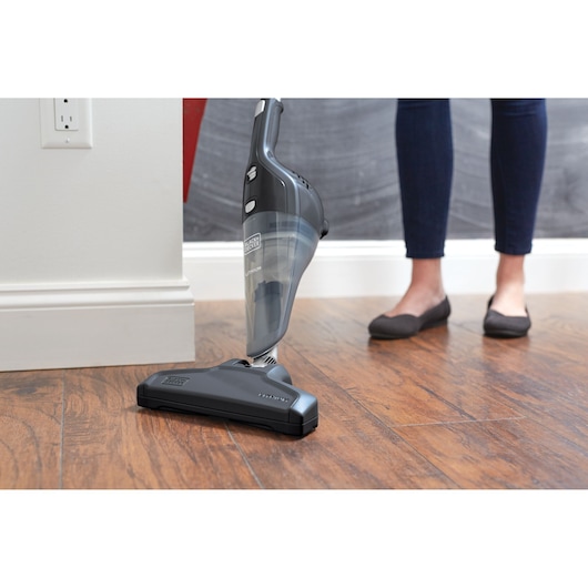 A BLACK+DECKER cordless stick vacuum cleaner is being used on a wooden floor, with a person standing nearby wearing dark jeans and black shoes.