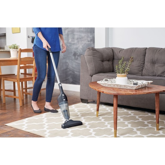 A person is using a BLACK+DECKER stick vacuum cleaner (model NSVA315J) to clean a patterned rug in a living room. The room features a sofa, a wooden coffee table with a decorative tray and a potted plant, and a dining area in the background.