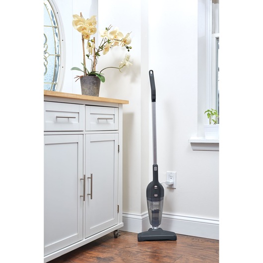 A BLACK+DECKER stick vacuum cleaner model NSVA315J stands upright against a white wall in a modern home. The vacuum is placed beside a white cabinet with a wooden top and a potted orchid, near a window with a small plant on the sill.