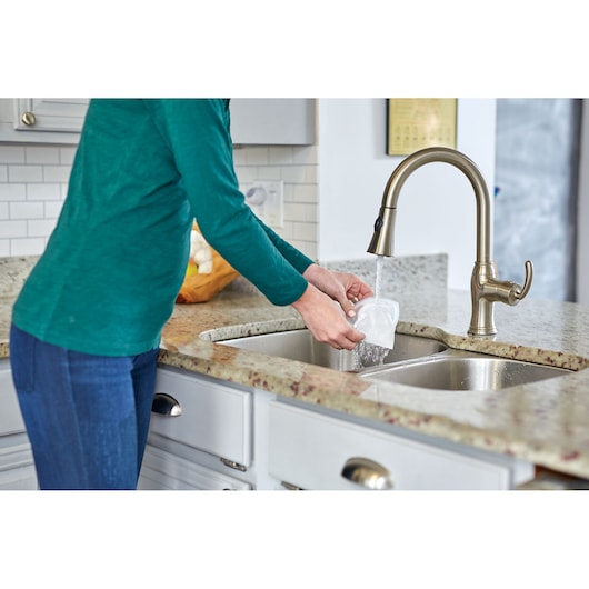 A person is rinsing a plastic container under a kitchen faucet at a sink with granite countertops.