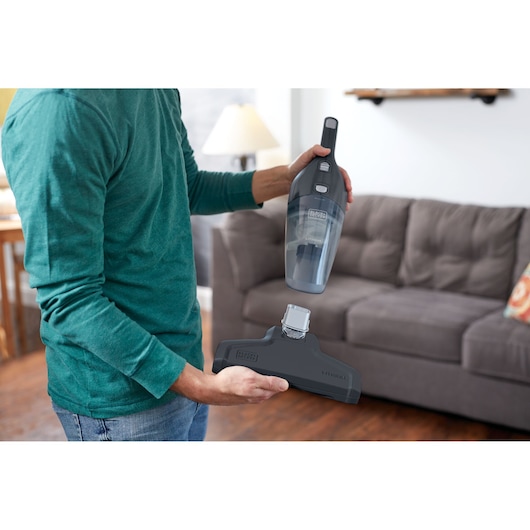A person is holding a BLACK+DECKER handheld vacuum cleaner (model NSVA315J) and its detachable floor attachment in a living room, with a sofa and lamp in the background.