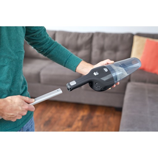 A person holds a BLACK+DECKER handheld vacuum cleaner and a nozzle attachment in a living room, with a grey sofa and colorful cushions in the background.