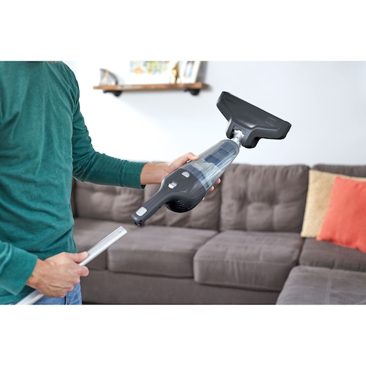 A person holding a BLACK+DECKER handheld vacuum cleaner in a living room, preparing to assemble it with a long extension tube. The vacuum is positioned near a brown sofa with orange and yellow pillows.