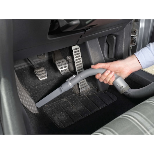 A person uses a handheld vacuum cleaner to clean the floor area near the pedals inside a car.