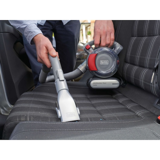 A person uses a BLACK+DECKER Dustbuster Flexi Auto handheld vacuum cleaner to clean the seat of a car. The vacuum is grey and red and has a flexible hose attachment for reaching tight spaces.