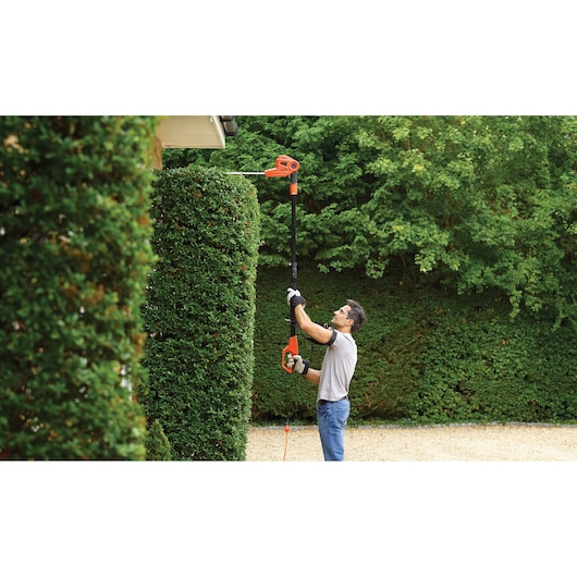 A person using a BLACK+DECKER pole hedge trimmer to cut tall hedges in a garden. The person is wearing gloves and safety gear, and the hedge trimmer is extended to reach the top of the hedge.