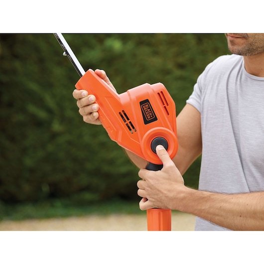 A person operating a BLACK+DECKER orange hedge trimmer outdoors, holding and adjusting the tool's handle.