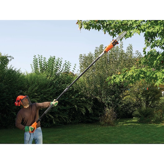 A person wearing protective gear uses a BLACK+DECKER PS7525 pole saw to trim branches from a tree in a garden.
