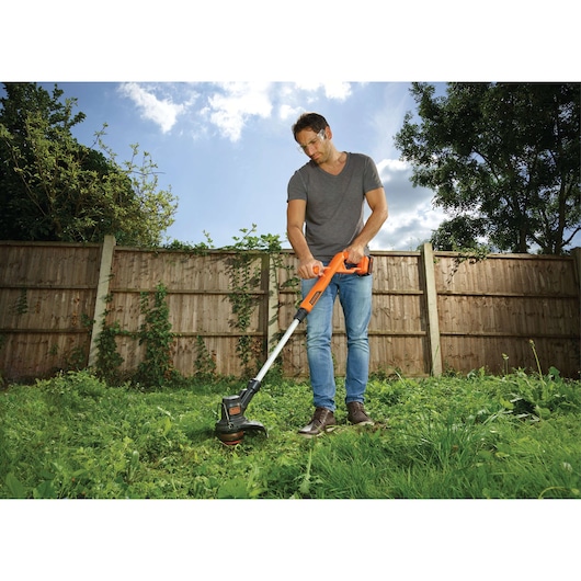 A person using an orange and black BLACK+DECKER grass trimmer in a garden with overgrown grass, standing near a wooden fence, with a clear sky above.