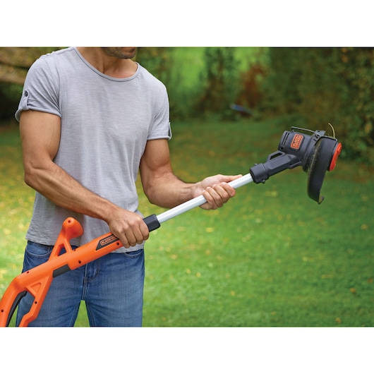 A person holding a BLACK+DECKER cordless grass trimmer in a garden, with a green lawn and bushes in the background.