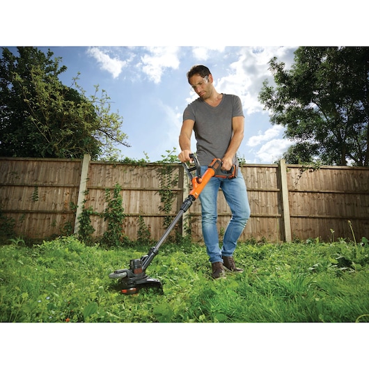 A person using a BLACK+DECKER cordless grass trimmer in a garden with tall grass, standing near a wooden fence under a partly cloudy sky.