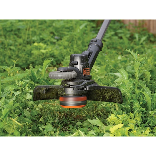 Close-up view of a BLACK+DECKER cordless grass trimmer cutting through thick green foliage outdoors.