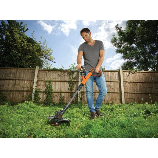 A person using a BLACK+DECKER STC1840PC cordless grass trimmer in a garden, standing on grass with a wooden fence and trees in the background. The person's face is blurred.