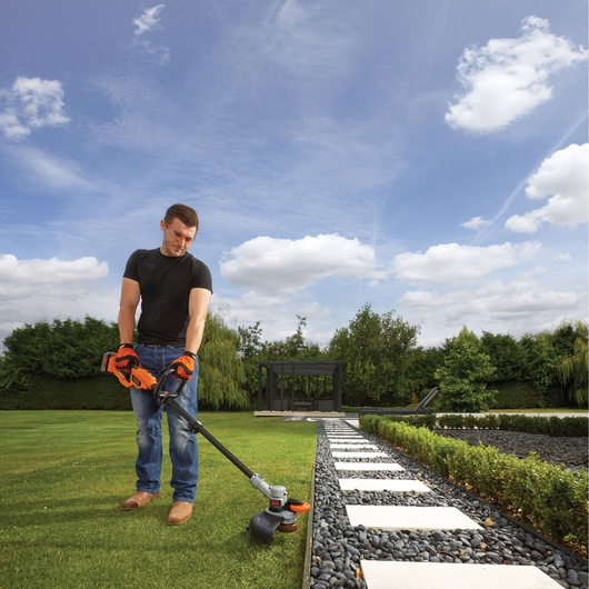 A person with a blurred face is using a BLACK+DECKER STC1840PC cordless grass trimmer to edge a garden lawn next to a stone pathway and landscaped area, under a partly cloudy sky.
