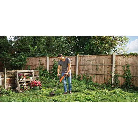 A person using a BLACK+DECKER cordless grass trimmer in a garden with wooden fencing, surrounded by plants and flowers. The person's face is blurred.