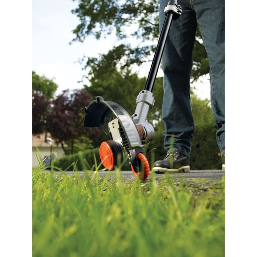 A close-up view of a person using a BLACK+DECKER cordless grass trimmer on the edge of a lawn, with green grass in the foreground and trees in the background.