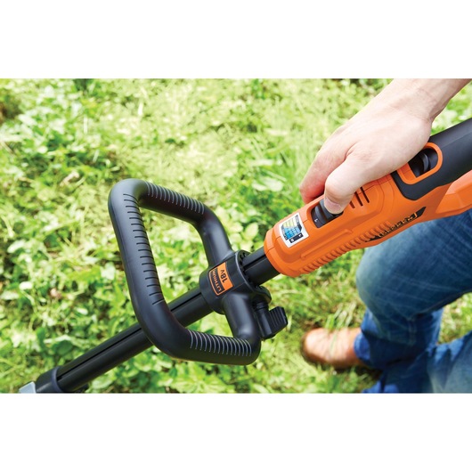 Close-up of a person using a BLACK+DECKER orange and black cordless grass trimmer outdoors on green grass. The person's hand is adjusting the handle.