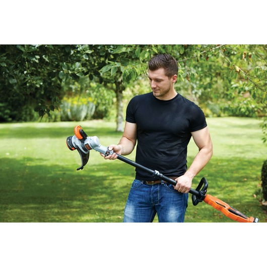 A person in a black t-shirt and jeans standing outside in a garden, holding a BLACK+DECKER STC1840PC cordless grass trimmer. The background shows green grass and trees, indicating an outdoor setting.